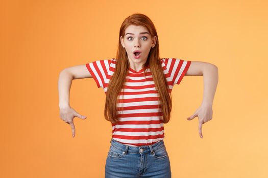 Shocked excited attractive redhead woman gasping, drop jaw shook, stare camera astonished, pointing down, feel amazed and surprised see incredible good offer, gossiping stunned, orange background.