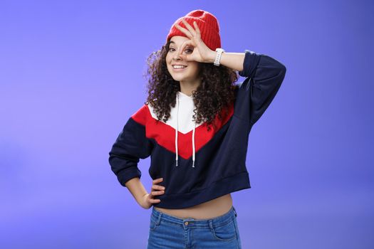 Okay girl agrees. Friendly-looking happy and charming young curly-haired girl in warm beanie showing ok gesture or circle over eye as peeking at camera and smiling broadly with joy over blue wall.
