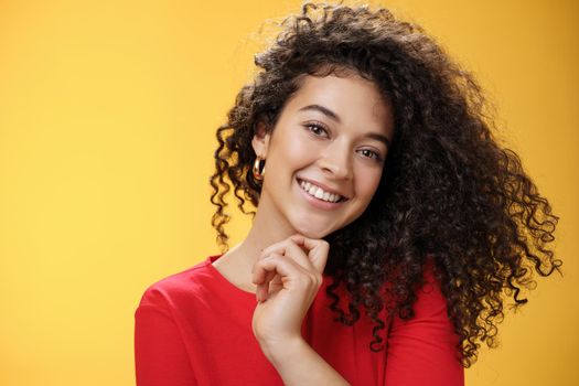 Close-up shot of stylish and happy bright curly-haired female in red dress tilting head sensually touching chin with finger and smiling broadly making flirty gazed at camera over yellow background.