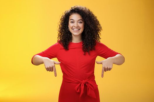 Studio shot of pleasant smiling young curly-haired woman in red dress pointing down with index fingers as promoting awesome event smiling broadly suggesting try or buy over yellow wall.