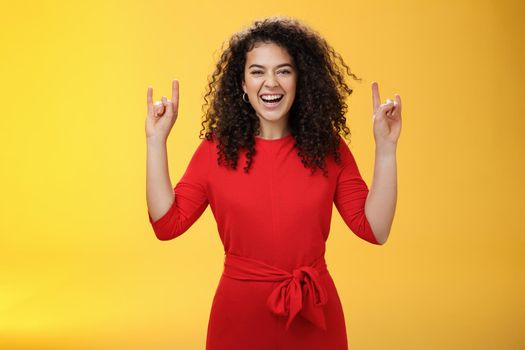 Rock n roll live. Charismatic excited young curly-haired woman in red dress feeling awesome as waiting concert of favorite band showing rock gestures with raised hands and smiling thrilled and happy.