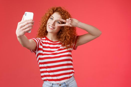 Hey followers how your summer holidays. Cheerful self-assured stylish fashionable redhead curly girl strike pose show peace victory gesture look smartphone screen taking selfie red background.