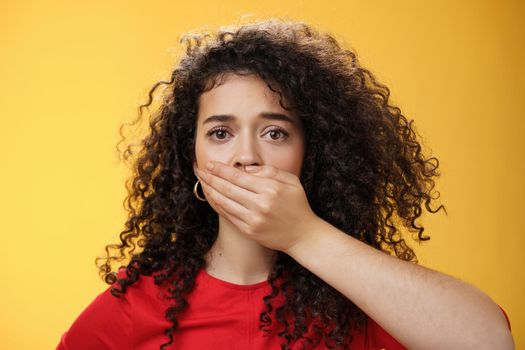 Close-up shot of concerned and insecure troubled woman keep silent scare of telling anyone her problem covering mouth with palm not to scream or slip word looking sad and sorrow over yellow background.