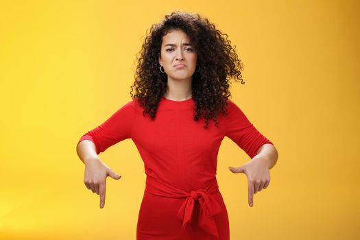 Gloomy disappointed cute silly curly-haired female in red dress frowning upset and making sad smile as pointing down with regret and unsatisfied expression, jealous or displeased over yellow wall.