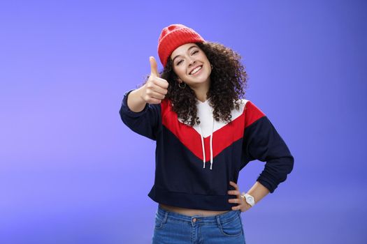 You doing great, like. Portrait of happy delighted young stylish woman in winter beanie and sweatshirt showing thumbs up pleased and satisfied approving and being supportive smiling at camera.