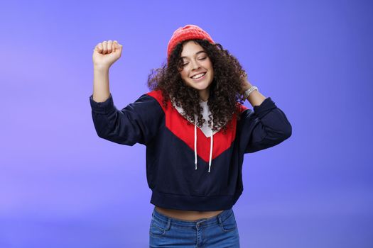 Girl delighted and carefree dancing over blue background with closed eyes and tender smile moving raised hands in rhythm of music, dreaming having positive mood wearing warm winter hat and sweatshirt.