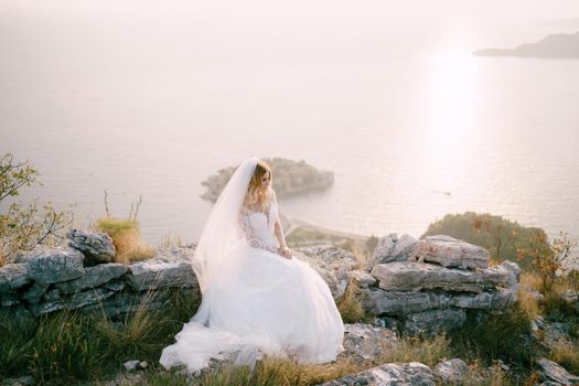 Bride in a white dress sits on a hill on the stones against the backdrop of the Sveti Stefan island. Montenegro. High quality photo