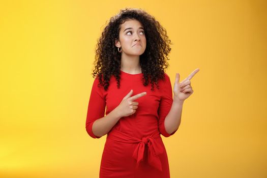 Girl pointing and looking at upper right corner with regret and sorrow missing good chance or opportunity getting upset and sad pulling gloomy smile with lips pursed down standing over yellow wall.