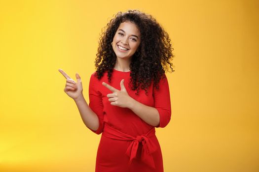 Friendly cute assertive female assistant in elegant red dress with curly hair tilting head flirty and smiling broadly, satisfied as pointing at upper left corner, recommending product over yellow wall.