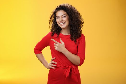 Portrait of friendly and assertive charming female coworker showing around newbie pointing left and smiling, laughing at camera having fun posing with hand on hip over yellow background. Advertisement concept