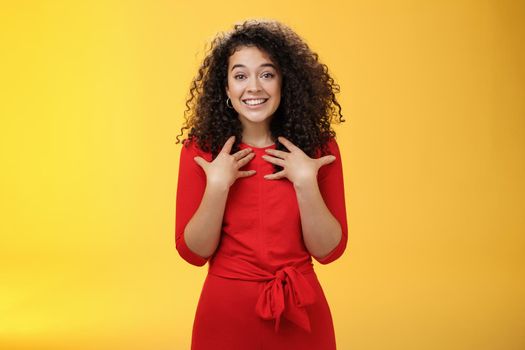 Pleased and thankful tender curly-haired kind girl in red dress pressing hands to breast being surprised with unexpected tender heartwarming gift thanking smiling delighted over yellow background
