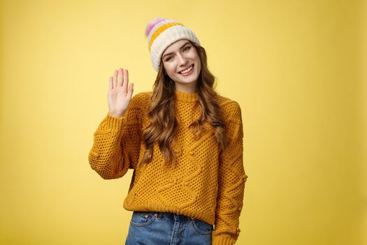 Friendly charming smiling young woman tilting head outgoing gaze waving hand hello hi gesture greeting you hey what up, standing carefree joyfully looking camera, yellow background.