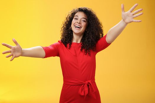 Come into my arms. Portrait of friendly and loving, caring charming woman with curly hair in red casual dress spread hands as wanting give hug smiling broadly at camera giving warm welcome or cuddle.