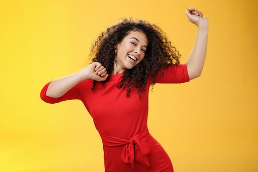Carefree girl feeling uplifted and joyful dancing in red dress raising hands up happily tilting head and smiling broadly at camera as enjoying vacation, celebrating holidays over yellow background.