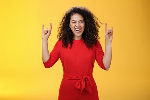 Cute excied and daring woman in 25s sticking out tongue as fooling around having fun on concert winking joyfully and showing rock-n-roll gesture feeling amused and alive over yellow background.