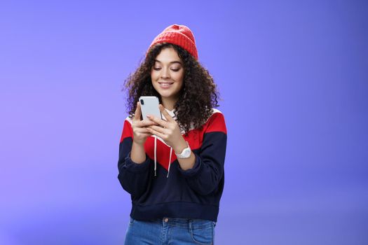 Woman searching in internet funny memes, holding mobile phone in hands smiling joyfully and cute at smartphone screen, typing message or browsing internet over blue background, wearing warm winter hat.