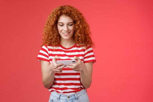 Redhead girl fool around waiting queue dentist playing awesome smartphone game hold phone horizontal tap cellphone screen look telephone display smiling delighted entertained standing red background.