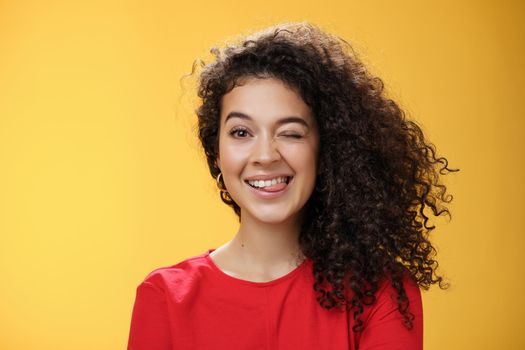 Portrait of funny and cool sister with curly hair winking playfully having fun and foolind around showing tongue as playing with siblings adoring spend time with chidren over yellow background.