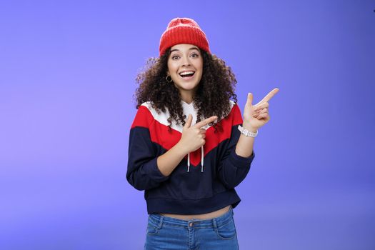 Enthusiastic friendly-looking attractive young 20s woman with curly hair in warm beanie and sweatshirt smiling open mouth amazed and delighted as pointing at upper right corner over blue background.