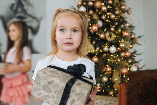 Very nice charming little girl blonde holds a gift on a background of Christmas trees in the interior of the house