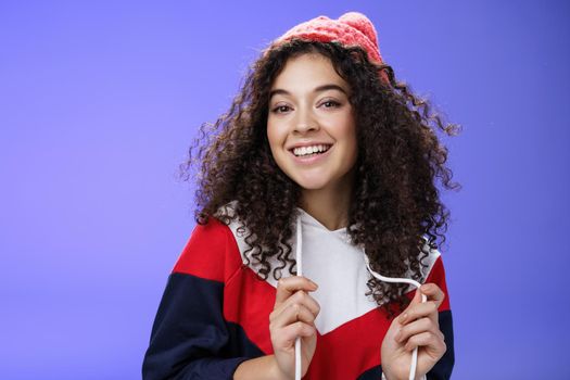 Close-up shot of carefree attractive feminine girl with curly hair in beanie playing with sweatshirt as posing over blue background, smiling at camera having fun and positive attitude.