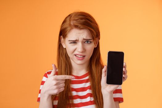 Perplexed disappointed redhead girl show smartphone screen app, pointing mobile display unsure, full disbelief, grimace doubtful, have hesitations, disapprove bad online choice, orange background.