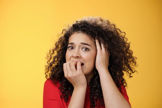 Scared woman overthinking frightened being in trouble biting fingers holding hand on head in despair, standing concerned, panicking and feeling uneasy as standing over yellow background. Emotions, facial expressions concept