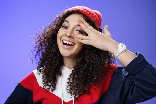 Close-up shot of charismatic happy carefree woman in winter red cute beanie and sweatshirt holding fingers near eye and peeking with broad smile at camera having fun, playing over blue background.