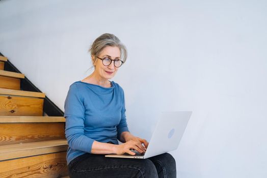 Elderly woman working at laptop while sitting on the steps at home