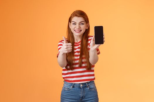 Happy satisfied redhead attractive girl cheering show thumb-up, recommend smartphone app, show mobile screen, introduce device, approve good dress show online shop cellphone, orange background.