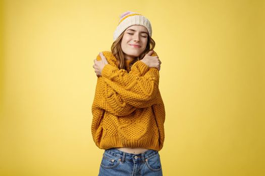 Upbeat charming cheerful young woman feeling lucky happy embracing herself like new warm comfortable sweater tilting head shoulder close eyes smiling broadly hugging own body, yellow background.