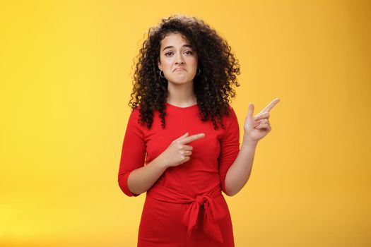 Waist-up shot of upset displeased girl with curly hair being down in dumps pointing at upper right corner with disappointed gloomy smile, feeling regret, sadness standing like loser over yellow wall.