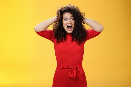 Girl feeling pressure standing anxious in panic holding hands on curly hair yelling at camera disturbed, freaked out being tensed and upset with bad situation, standing troubled over yellow background.