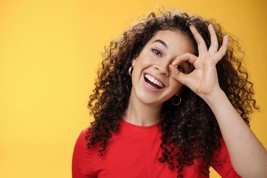 Hey I see you. Charismatic funny and playful cute tender girlfriend with curly hair showing okay or zero gesture over eye peeking at camera and smiling broadly, tilting head right over yellow wall.