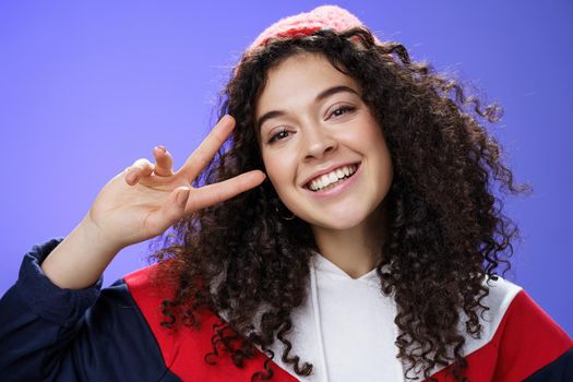 Close-up shot of friendly charming curly-haired woman with warm beanie tilting head joyfully smiling broadly showing victory or peace gesture near face, feeling happy and delighted over blue wall.