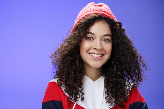 Headshot of tender and cute curly-haired 20s woman in warm beanie and cool sweatshirt smiling broadly enjoying awesome sunny and chilly days outdoors having fun posing over blue background. Copy space