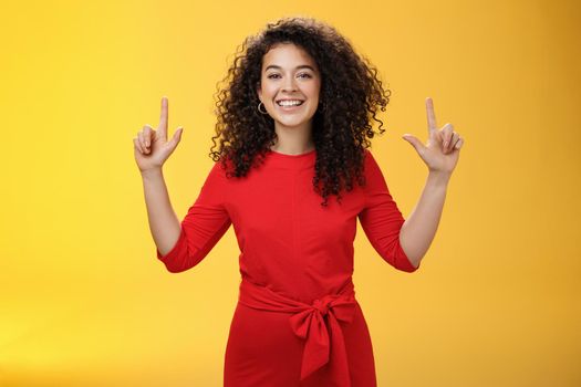 Carefree joyful and friendly successful woman with curly hairstyle in red dress smiling delighted and ambitious as pointing up with raised hands giving recommendation or showing promotion.