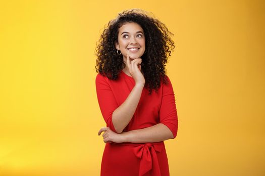 Girl having interesting idea, smiling as feeling confident plan might work. Charming happy woman with curly hair in red dress gazing at upper left corner thoughtful, thinking over yellow wall.