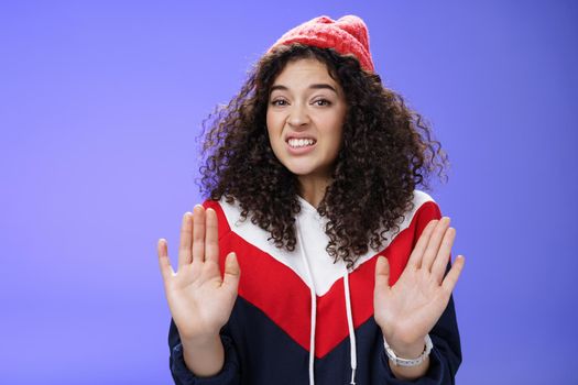 Displeased european woman with curly hair raising palms near chest and waving in refusal and rejection gesture grimacing unsatisfied and unwilling to participate in suspicious action over blue wall.