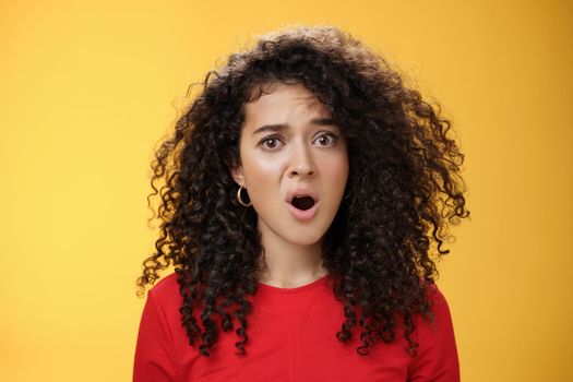 Confused and frustrated young questioned woman with curly hair open mouth and raising eyebrow in surprise being displeased with unfair situation standing clueless and upset over yellow background.