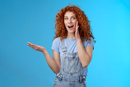 Amused excited attractive cheerful redhead curly girl present awesome product hold raised hand blank blue copy space share secret promo offer palm near opened mouth studio background.