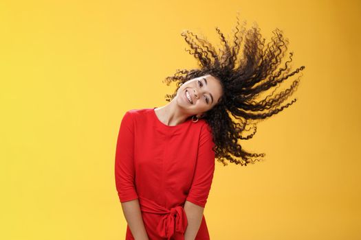 Bright happy and carefree playful woman waving curly hair making wave and smiling broadly as standing joyful in red dress over yellow background in good mood for future adventures. Copy space
