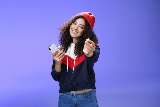 Wanna listen music with me. Portrait of friendly charming and cute woman in warm beanie extending arm towards camera to suggest take wireless earphone and enjoy song together, holding smartphone.