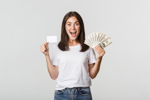 Excited happy brunette girl showing credit card and money, smiling over white background.