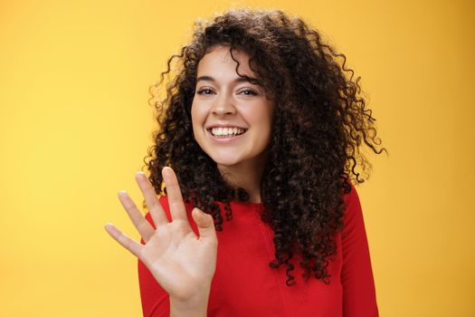 Charming friendly and self-assured attractive curly woman waving cute with palm to say hi or hello smiling broadly greeting man trying flirt in party posing joyful over yellow background.
