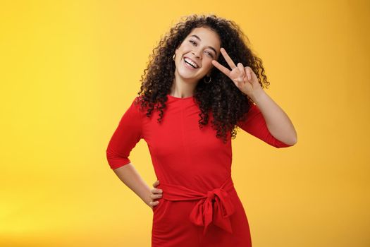 Waist-up shot of friendly tender and feminine cute woman with curly hairstyle tilting head and smiling, laughing and showing victory or peace gesture at camera over yellow background.