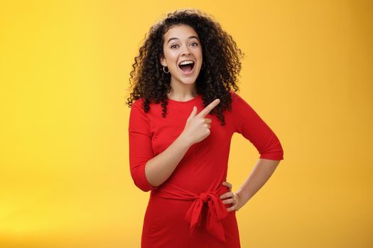 Waist-up shot of amused and impressed beautiful caucasian girl with curly hair in red dress open mouth with amazement and joy and pointing at upper right corner checking out awesome copy space.