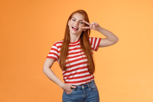 Friendly attractive modern redhead female college star show peace victory sign near eye, smiling happily, express positivity, joy and confidence, motivated win, stand orange background optimistic.