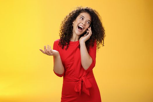 Talkative glamour silly girl with curly hair having fun feeling carefree and happy taling on mobile phone turning away as laughing joyfully gesturing with hand holding smartphone pressed to ear.