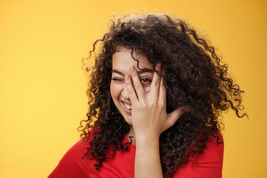 Close-up shot of sensual and playful attractive girlfriend with curly hair covering face with palm and peeking through fingers with happy tender smile anticipating surprise over yellow background.